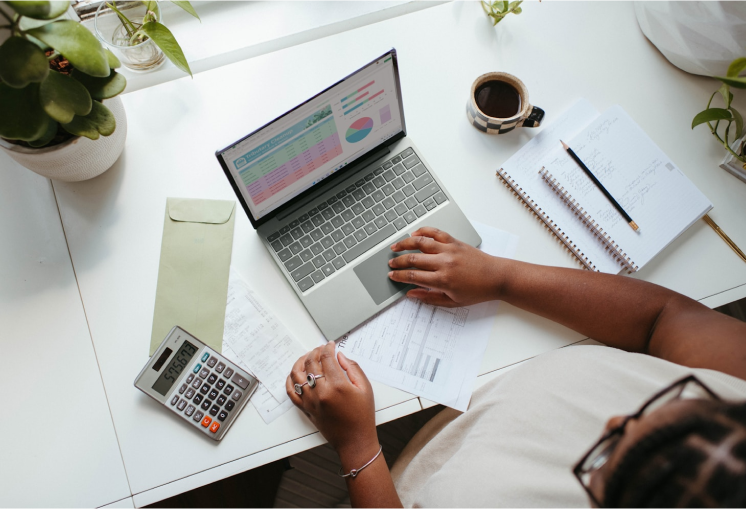 Pessoa trabalhando em uma mesa de escritório, utilizando um notebook com gráficos e dados exibidos na tela. Há uma calculadora, anotações em papel, um envelope e uma xícara de café ao lado. Ambiente iluminado e organizado com plantas ao fundo.